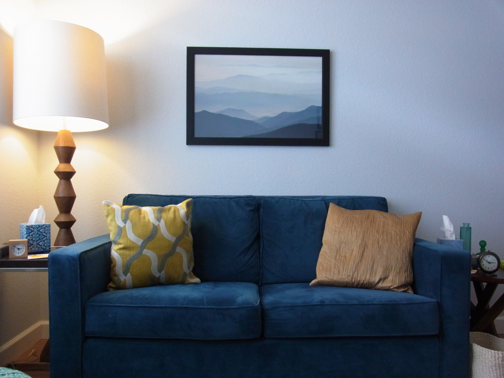 A blue loveseat with two yellow pillows sits below a framed photo of a blue mountain range.  Counseling south Austin offers affordable EMDR therapy and counseling services in south Austin.